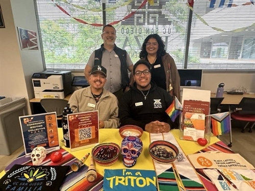 A group of four people is pictured inside a brightly decorated room, smiling and seated at a colorful table. Two individuals are seated, and two are standing behind them. The table is adorned with vibrant decorations, including colorful bowls, sugar skulls, a "I'm a Triton" towel, and brochures for UC San Diego events and programs. Rainbow flags and other cultural symbols are visible on the table, indicating inclusivity and celebration of diversity. A large window in the background shows part of the UC San Diego logo.